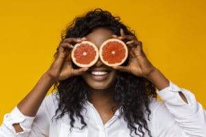 fille rigolote qui se met deux demi pamplemousses ouvert en guise de lunettes, devant ses yeux sur fond jaune.